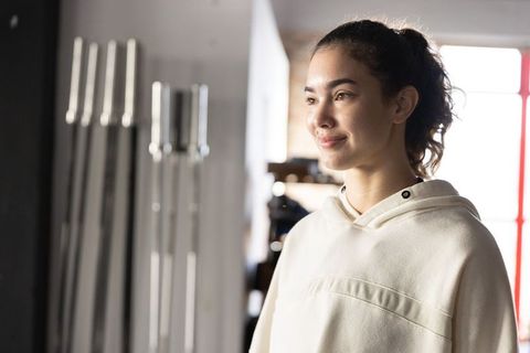Young woman in hoodie smiling after workout in light-filled gym with weight bars