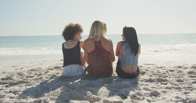 Three friends are sitting on sandy beach with ocean in background, enjoying warm, sunny day. They are seen from behind, talking and bonding. Dual shade tops enhance beach vibes. Ideal for vacation, summer, friendship, relaxation themes, and travel promotions.