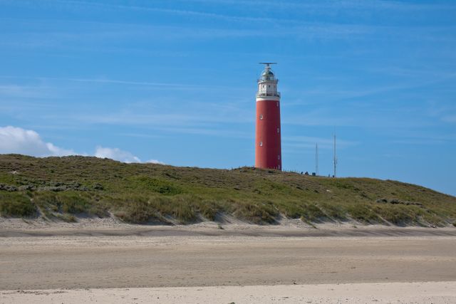 Red lighthouse standing on sandy beach with blue sky in background. Perfect for travel brochures, vacation advertisements, and coastal landscape scenes.