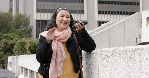 Mature woman waving while using smartphone voice message on urban plaza