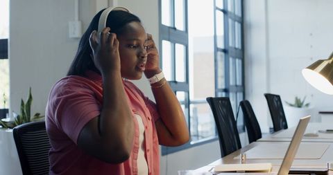 African woman adjusting white headphones while preparing workspace at modern coworking desk