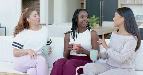 Three friends chatting and sipping mugs on couch in cozy living room loungewear
