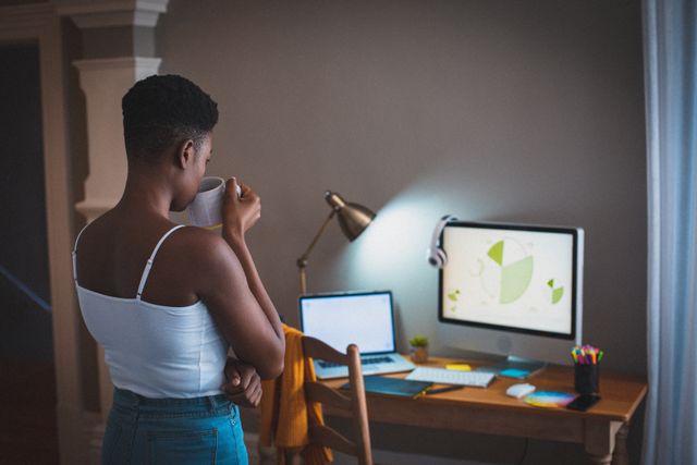 African American woman standing at home office desk, drinking coffee while looking at computer screen. Ideal for illustrating remote work, home office setups, productivity during quarantine, and modern lifestyle. Suitable for articles, blogs, and advertisements related to working from home, freelance work, and technology use.
