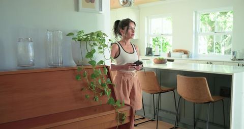 Indian woman standing in modern kitchen holding smartphone near piano, bright lifestyle moment