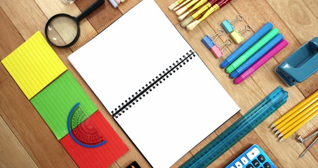 A variety of colorful school supplies are neatly arranged around a blank notebook on a wooden surface, with copy space. The setup suggests a preparation for educational activities or a back-to-school theme.