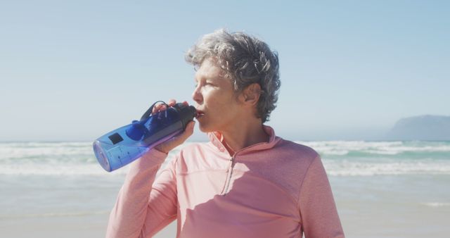Senior caucasian woman wearing sports clothes and drinking water at beach. Retirement, free time, healthy and active lifestyle.