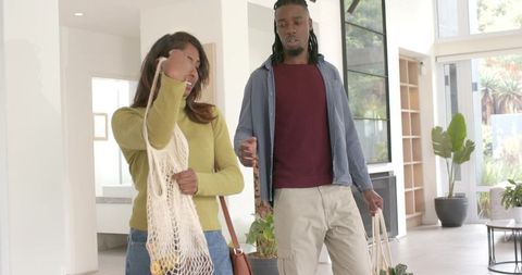 Diverse couple entering modern home carrying reusable bags and potted plant, eco living