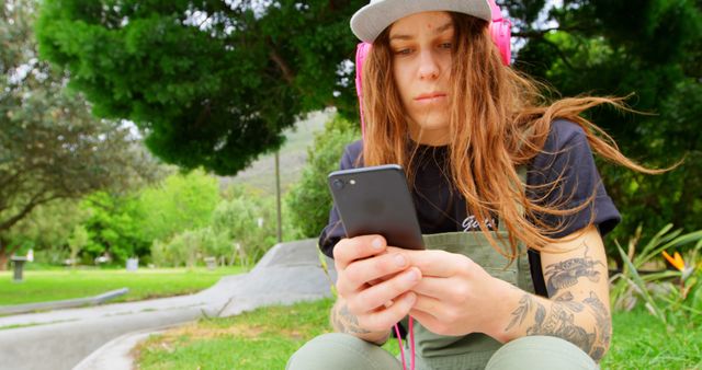 A young woman with tattoos is sitting outdoors in a park, concentrating on her smartphone while wearing pink headphones. The woman, dressed casually in overalls and a t-shirt, exudes a laid-back and modern vibe. This image can be used to depict themes of modern lifestyle, technology use, casual fashion, and outdoor activities.