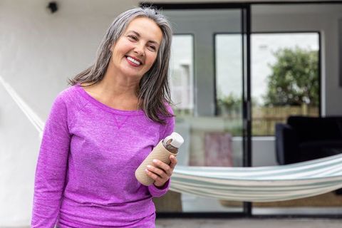Smiling mature woman holding water bottle on sunny patio with hammock and plants