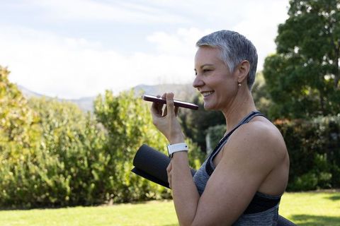 Mature woman with short gray hair speaking into smartphone holding yoga mat outdoors