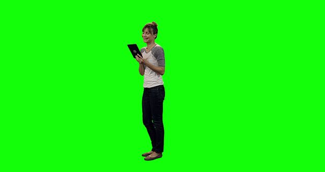 Woman standing against a green screen background using a tablet device. She is casually dressed in a shirt and jeans, appearing focused on the screen. Ideal for ads or content depicting technology usage, app promotion, or a digital lifestyle. The green screen can be replaced with any background.