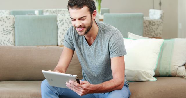 A young Caucasian man is seated on a sofa, engaging with a tablet with a smile, with copy space - Download Free Stock Photos Pikwizard.com