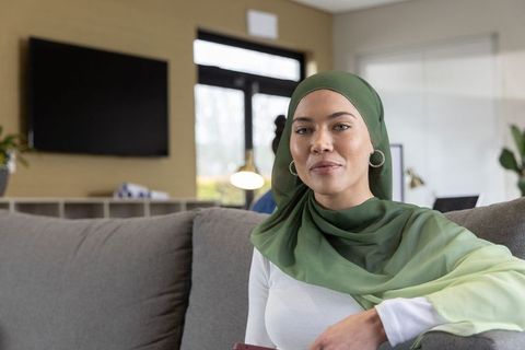 Confident muslim woman in green hijab sitting on gray sofa holding phone in office lounge