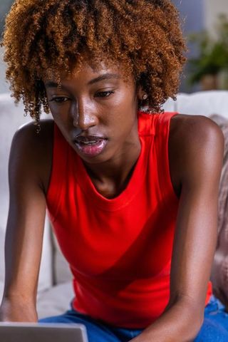 Young african american woman working on laptop from couch wearing red top and jeans