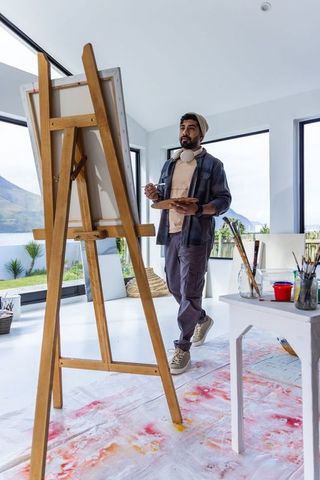 Asian male artist painting on canvas in sunlit minimalist studio with lakeside views and workspace