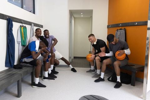 Multiracial basketball teammates laughing and preparing on locker room benches, team camaraderie