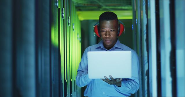 An engineer in a data center is monitoring systems using a laptop. He is wearing earmuffs for noise protection and standing between server racks under cool lighting. Ideal for content related to IT, data management, network support, or technology infrastructure maintenance.