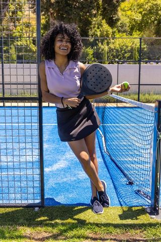 Smiling african american woman leaning on padel court gate holding paddle and ball