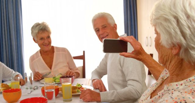 Seniors sit at dining table with plates of food, smiling and enjoying each other's company while one takes a picture using a smartphone. Ideal for themes related to senior living, friendship, social gathering, and healthy lifestyle.