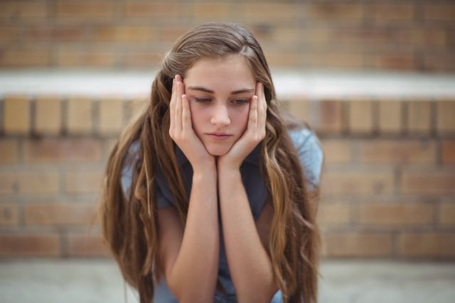 Teenage girl sitting alone against a brick wall, looking sad and thoughtful. Ideal for use in articles or campaigns related to mental health, teenage depression, loneliness, school life, and emotional well-being.