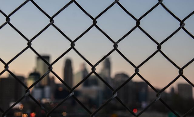Image shows a view of a downtown city skyline through a chain-link fence during sunset, with buildings blurred in the background. Can be used to represent themes of urban life, security, confinement, or the contrast between natural and industrial elements. Ideal for blog posts, urban studies, social issues, or travel articles.