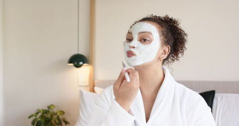 African american woman applying clay face mask in robe on bed morning skincare routine