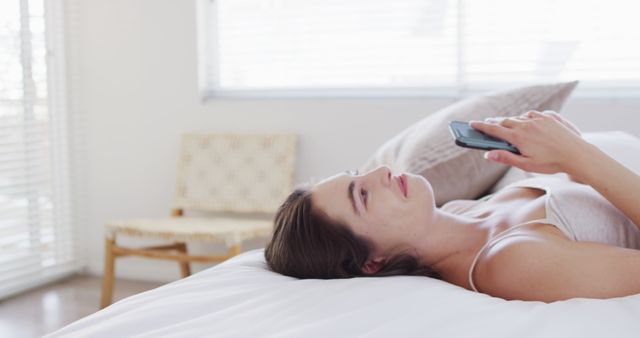 Woman lying on a white bed, using a smartphone, appearing casual and relaxed. The background shows a modern bedroom with soft lighting and minimal decor. Perfect for content related to lifestyle, technology use, relaxation, home living, and personal well-being. Ideal for articles or advertisements on self-care, leisure activities, or home design inspiration.