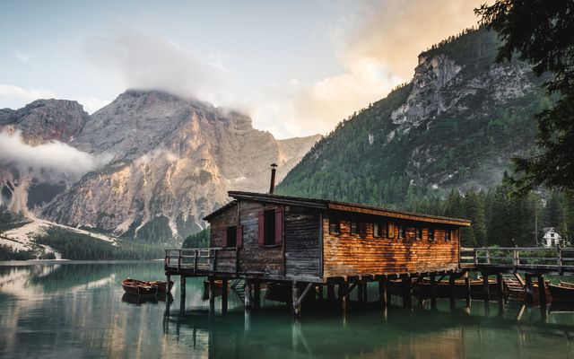 Scenic view of a wooden dock house on a tranquil mountain lake at sunrise. Reflection of the mountains and house in the calm water adds to the serene ambiance. This image captures the peace and beauty of untouched nature, ideal for travel and adventure promotions, nature conservation campaigns, and creating a calming, picturesque atmosphere for websites and print media.