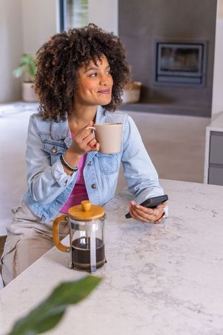 African american woman enjoying coffee at kitchen island holding mug and phone