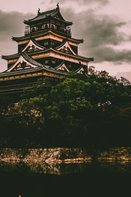 Traditional Japanese castle illuminated by sunset, showcasing its intricate design and historical significance. The dark clouds add a dramatic effect, enhancing the mystical atmosphere. The serene moat with reflections adds to the beauty of the composition. Perfect for travel guides, architecture magazines, cultural studies, and historical documentaries.