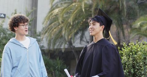 Recent graduate celebrating with friend on campus wearing cap gown holding diploma