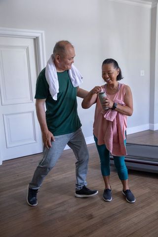 Asian mature couple sharing water bottle and towels in home gym after workout session