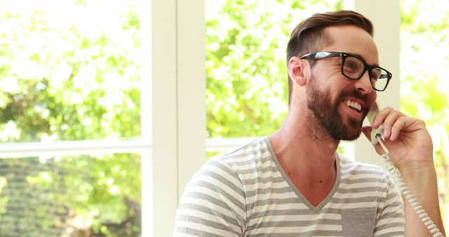 Smiling Young Man Talking on Corded Phone Sitting Near Window - Download Free Stock Images Pikwizard.com