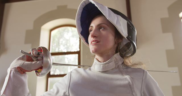 This image features a female fencer holding an épée sword in a sunlit indoor room. She is wearing full protective fencing gear and exhibits a determined expression, ready for a match or sparring session. Such an image is ideal for use in promotional materials for fencing events, sports training programs, motivational posters, and articles about women's participation in competitive sports.