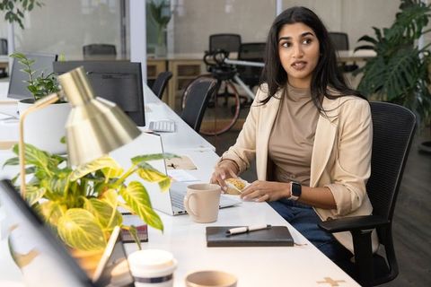 Indian woman enjoying pastry at coworking desk with laptop and coffee