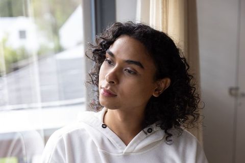 Contemplative non-binary adult gazing through window in white hoodie, soft natural light