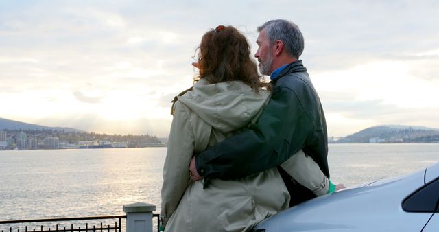 A middle-aged Caucasian couple enjoys a scenic view of a waterfront at sunset, with copy space. Their embrace suggests a moment of shared tranquility as they overlook the serene landscape.