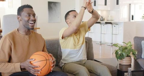 Two friends cheering while watching basketball game at home holding basketball on couch