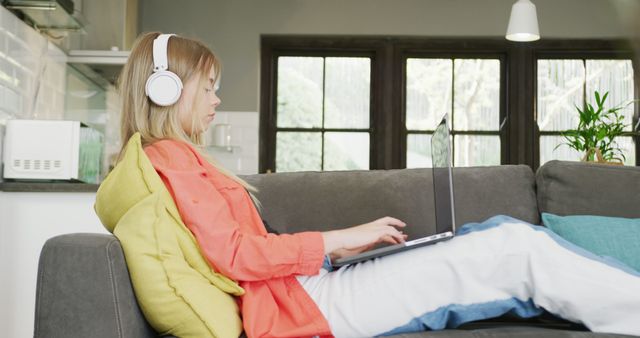 Teen girl wearing headphones while using a laptop on a comfortable sofa, exemplifying modern lifestyle. Perfect for content on online learning, remote work, technology for teens, and relaxed home environments.
