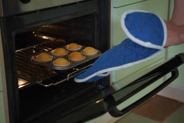 This image shows a woman placing a tray of muffins into an oven in a home kitchen. She is wearing blue oven mitts for safety. This image can be used for articles or advertisements related to home baking, cooking tutorials, kitchen safety, or lifestyle blogs focusing on homemade desserts and culinary activities.