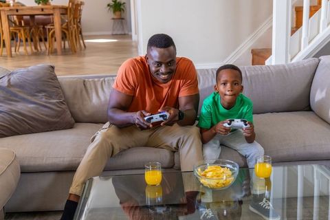 African American dad and son gaming on sofa with snacks and juice