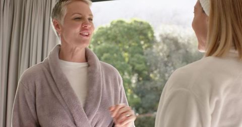 Two women sharing cozy morning conversation in plush robes by sunlit window