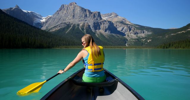 Woman in bright life jacket seen paddling in a canoe on a calm turquoise lake, flanked by tall mountains with snowy peaks. Perfect image for concepts related to outdoor adventure, summer recreation, travel, and serenity. Ideal for travel brochures, adventure trip promotions, fitness advertisements, and mindfulness or relaxation products.