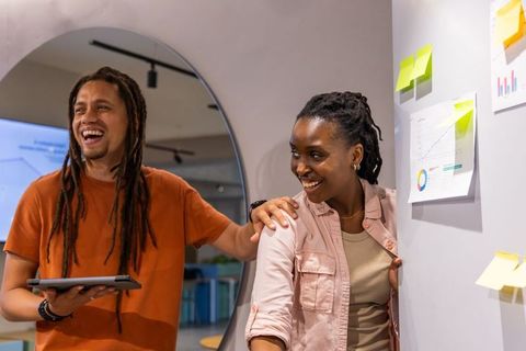 Diverse team laughing and brainstorming by chart wall in modern office with tablet and sticky notes