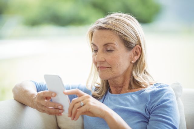 Senior woman sitting on couch using mobile phone in living room. Ideal for depicting technology use among elderly, modern lifestyle, communication, and home comfort. Suitable for articles on senior technology adoption, mobile communication, and home living.