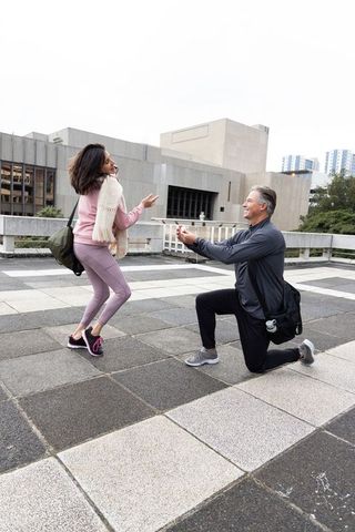 Man proposing on rooftop terrace while woman reacting with surprise in urban plaza cityscape