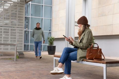 Urban woman sitting on bench using smartphone while man walking by in modern plaza