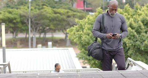 Senior african american man walking up park stairs in athletic gear checking smartphone