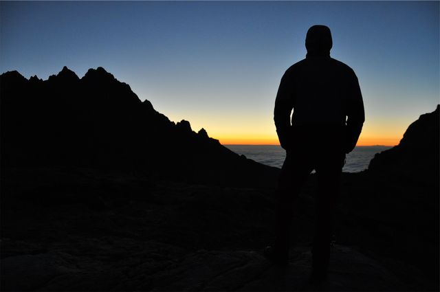 Silhouette of a hiker standing on terrain, watching sunrise over a mountain ridge. Ideal for themes of adventure, nature, travel, and personal growth. Suitable for outdoor activity promotions, inspirational content, or landscape photography portfolios.