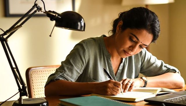 Writing indian woman focusing at desk with notebook and lamp for remote work and study